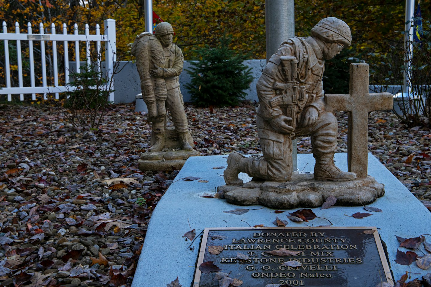 Stone soldier statues stand in autumn leaves at a memorial.